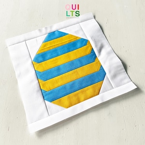 May include: A square quilt block with a geometric design. The centre features a yellow and blue striped pattern, surrounded by a white border. The text "QUILTS AND NICE STUFF" is in the upper right corner.