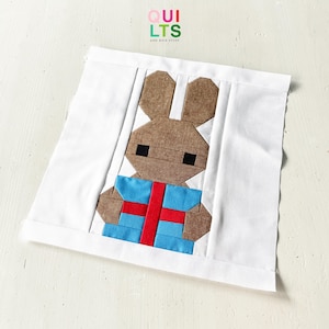 May include: A white square fabric featuring a brown bunny holding a blue gift with a red ribbon. The bunny has black square eyes and is made of geometric shapes. The text "QUILTS AND NICE STUFF" is in the upper left corner.