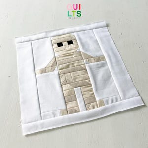 Op de afbeelding: Een handgemaakt quiltblok met een mummie-ontwerp. De mummie is gemaakt van beige stoffen stroken met zwarte vierkante ogen, tegen een witte achtergrond. Het blok is omlijst met witte stof en de tekst "Quilts and Nice Stuff" is zichtbaar.