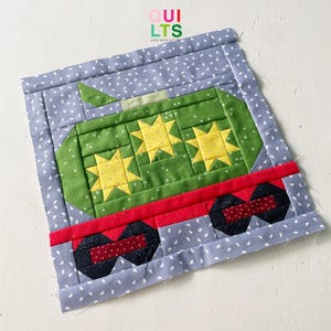 Op de afbeelding: Een groene en gele patchwork quiltblok met een trein met drie gele sterren. De trein staat op een rode en zwarte achtergrond met een blauwe en witte gepatenteerde rand.