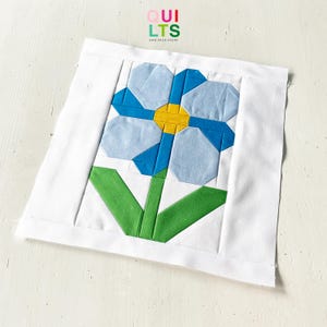 May include: A handmade quilt block with a blue flower design, yellow centre, and green stem and leaves, set against a white background. The text "QUILTS AND NICE STUFF" is in the upper right corner.