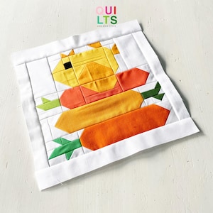May include: A patchwork quilt block depicting a yellow duck with orange and yellow carrots. The duck has a black eye and an orange shirt. The carrots are orange and yellow with green tops. The block is framed with white fabric. The text "QUILTS" and "AND NICE STUFF" is in the upper right corner.