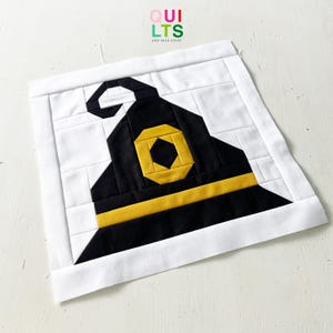 Könnte beinhalten: Ein Quiltblock mit einem schwarzen Hexenhut mit gelbem Band und Schnalle auf weißem Hintergrund. Der Hut ist der Mittelpunkt, mit dem Text "QUILTS AND NICE STUFF" in der oberen rechten Ecke.