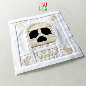 Könnte beinhalten: Ein quadratischer Stoffblock mit einem Mumien-Design, hergestellt aus verschiedenen Stoffstücken. Das Gesicht der Mumie ist beige mit schwarzen Augen und Mund. Der Block ist von weißem Stoff umrandet und enthält den Text "QUILTS AND NICE STUFF".