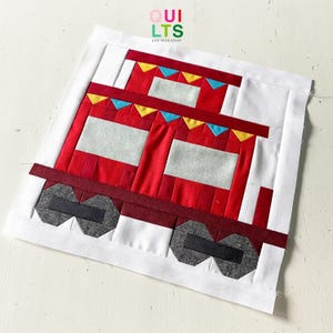 May include: A handmade quilt block featuring a red train with gray wheels and white windows. The train has decorative yellow and blue triangles along the top. The block is set against a white background. The text "QUILTS AND NICE STUFF" is in the top right corner.