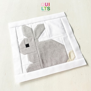 Può includere: Un blocco di tessuto patchwork con un disegno di coniglio grigio, un occhio nero e un piede beige. Il coniglio è posto su uno sfondo bianco e incorniciato da un bordo bianco. Il testo "QUILTS AND NICE STUFF" è nell'angolo in alto a sinistra.