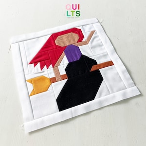Könnte beinhalten: Ein quadratisches Patchwork mit einer Hexe auf einem Besen. Die Hexe hat rotes Haar, einen lila Hut und ein schwarzes Kleid. Der Besen ist braun und gelb. Der Hintergrund ist weiß mit dem Text "QUILTS AND NICE STUFF" in Grün und Weiß.