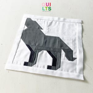 Könnte beinhalten: Ein handgefertigtes Quiltquadrat mit einem geometrischen Wolfsdesign in Grau und Schwarz auf weißem Hintergrund. Das Quadrat ist mit weißem Stoff eingefasst und enthält den Text "Quilts and Nice Stuff".