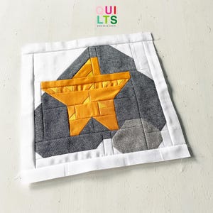 Könnte beinhalten: Ein gelber Stern-Patchworkblock auf grauem Hintergrund. Der Stern besteht aus kleineren Dreiecken und der Hintergrund aus Quadraten. Der Patchworkblock ist von einem weißen Rand umgeben.