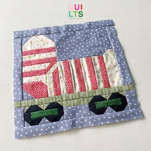 Peut inclure: Un bloc de courtepointe patchwork représentant un camion à benne basculante rouge, blanc et vert sur un fond bleu. Le camion a une caisse rayée et une benne à points. Le fond est un tissu bleu clair à pois blancs.