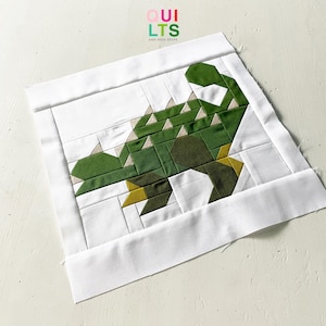 May include: A white fabric quilt block with a green and yellow dinosaur design. The dinosaur is made up of geometric shapes and is outlined in white. The quilt block is surrounded by a white border.