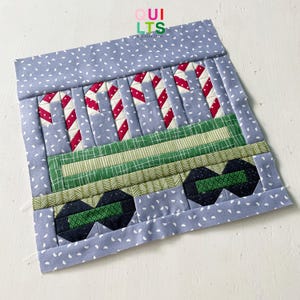 May include: A blue and white fabric block with a green and red candy cane design. The block is a quilted design with a train theme. The block is part of a larger quilt project.
