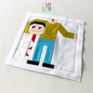 Könnte beinhalten: Ein handgefertigtes Quiltquadrat mit einer geometrischen Figur in einem weißen Rahmen. Die Figur hat ein hellbraunes Gesicht, schwarze Augen und grüne Haare. Sie trägt ein gelbes Hemd, eine blaue Hose und eine rote Krawatte. Der Text "QUILTS AND NICE STUFF" befindet sich oben rechts.