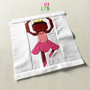 May include: A fabric square featuring a patchwork design of a ballerina. The ballerina is depicted in shades of pink, brown, and gold, with a crown. The background is white, and the text "QUILTS AND NICE STUFF" is visible.