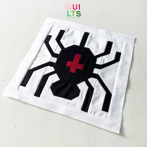 Könnte beinhalten: Ein weißer quadratischer Stoffblock mit einem schwarzen Spinnendesign und einem roten Kreuz. Die Spinne hat einen schwarzen Körper und Beine. Der Text "QUILTS AND NICE STUFF" befindet sich in der oberen linken Ecke.