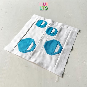 Puede incluir: Un cuadrado de tela blanca con cuatro hexágonos azules cosidos encima. La tela está doblada y los bordes están deshilachados. El texto "OUI LTS AND NICE STUFF" está impreso en la esquina superior izquierda.