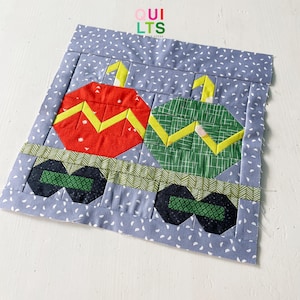 May include: A quilt block featuring two Christmas ornaments, one red and one green, with yellow zig-zag patterns. The ornaments are on a blue background with white dots. The block is surrounded by a green and black border.