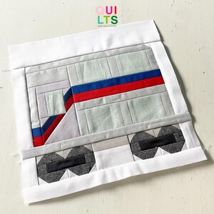 May include: A handmade quilt block featuring a geometric design of a train. The train is primarily gray with red and blue stripes. The wheels are black and gray. The block is framed with a white border. The text "QUILTS AND NICE STUFF" is in the upper right corner.