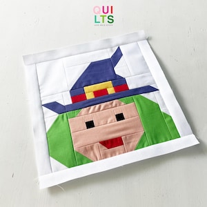 Peut inclure: Un bloc de patchwork coloré représentant une sorcière pixélisée. La sorcière a des cheveux verts, un chapeau bleu avec des accents rouges et jaunes, et un visage rose avec une bouche rouge et des yeux noirs. Le bloc est encadré d'une bordure blanche. Le texte "QUILTS AND NICE STUFF" est en haut à droite.