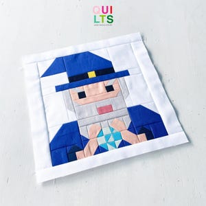 May include: A fabric quilt block featuring a pieced design of a wizard wearing a blue hat with a yellow band, a grey beard, and a blue robe. The wizard holds a light blue and white geometric crystal shape. The block has a white border.
