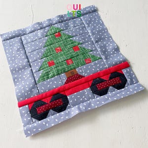 Op de afbeelding: Een blauwe en witte stoffen quiltblok met een groene patchwork kerstboom en een rode wagen met zwarte wielen. Het quiltblok is versierd met witte stippen.