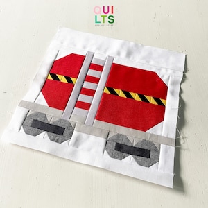 May include: A fabric block featuring a red and gray train car design with black wheels and yellow and black stripes. The design is set against a white background. The text "QUILTS and NICE STUFF" is in the upper right corner.
