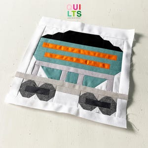 Könnte beinhalten: Ein handgefertigter Quiltblock mit einem Eisenbahnwaggon-Design. Der Block hat einen weißen Rand und verwendet Stoffe in Schwarz, Orange, Türkis und Grau. Der Text "QUILTS AND NICE STUFF" ist oben rechts sichtbar.