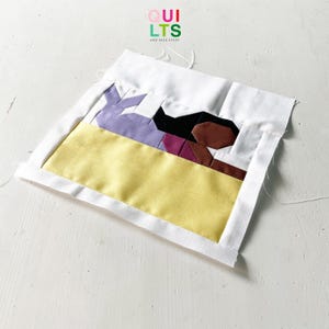 Op de afbeelding: Een quiltblok met een geometrisch ontwerp in geel, paars, bruin en zwart. Het blok is omgeven door een witte rand. De tekst "QUI LTS AND NICE STUFF" is zichtbaar in het midden bovenaan de afbeelding.