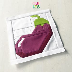 Könnte beinhalten: Ein Stoff-Quiltblock mit einem geometrischen Auberginen-Design. Die Aubergine ist lila mit einem dunkelroten Umriss und einem grünen Stiel. Der Block ist vor einem weißen Hintergrund mit einem weißen Rand angeordnet. Der Text "QUILTS AND NICE STUFF" befindet sich in der oberen rechten Ecke.