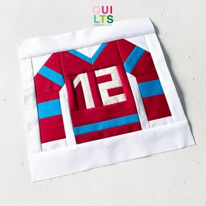 May include: A handmade quilt block featuring a red and blue jersey with the number 12 in white. The jersey has blue stripes and a white collar. The words "QUILTS AND NICE STUFF" are in the top right corner.