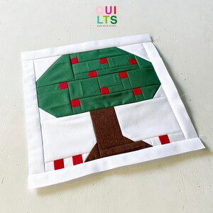 Puede incluir: Un bloque de edredón de tela cuadrado con un diseño de árbol hecho de piezas. El árbol tiene un tronco marrón y una copa verde con pequeños cuadrados rojos que representan frutos. El diseño está enmarcado por un borde blanco, con el texto QUILTS AND NICE STUFF arriba.