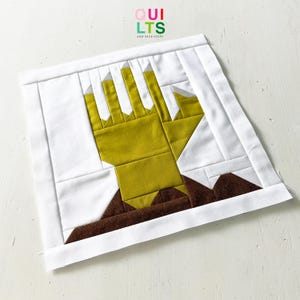 Könnte beinhalten: Ein quadratischer Quiltblock mit einem Handdesign in Olivgrün, Braun und Weiß. Die Hand ist der Mittelpunkt, mit detaillierten Finger- und Handflächenformen. Der Block ist mit weißem Stoff eingefasst, und der Text "QUILTS AND NICE STUFF" befindet sich in der oberen rechten Ecke.