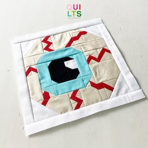 Könnte beinhalten: Ein quadratischer Stoffblock mit einem stilisierten Augendesign. Das Auge hat eine hellblaue Iris, eine schwarze Pupille und rote Adern. Der Block ist mit weißem Stoff umrandet. Der Text "QUILTS AND NICE STUFF" befindet sich in der oberen rechten Ecke.