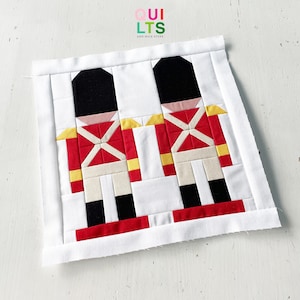 May include: Two red and white toy soldier figures with black hats and yellow accents on a white background. The figures are made of fabric and are sewn together. The background is a white fabric with a white border.