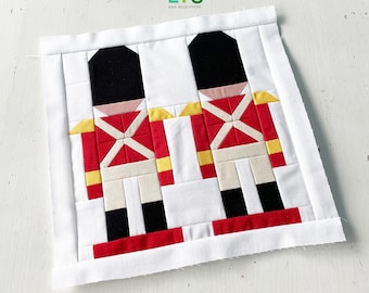 May include: Two red and white toy soldier figures with black hats and yellow accents on a white background. The figures are made of fabric and are sewn together. The background is a white fabric with a white border.