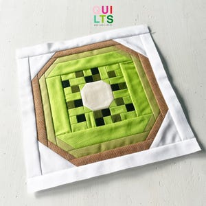 Könnte beinhalten: Ein quadratischer Quiltblock mit einem Kiwi-Frucht-Design. Die Mitte hat ein weißes Achteck, umgeben von Grüntönen und schwarzen Quadraten, mit einem braunen Rand und weißen Kanten. Der Text "QUILTS AND NICE STUFF" befindet sich in der oberen rechten Ecke.