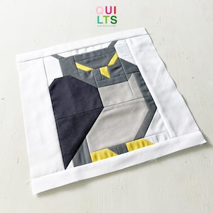 Könnte beinhalten: Ein quadratisches Stoffstück mit einem geometrischen Eulenmuster. Die Eule besteht aus grauen, schwarzen und gelben Stoffstücken, vor einem weißen Hintergrund. Der Text "QUILTS AND NICE STUFF" ist in der oberen linken Ecke sichtbar.