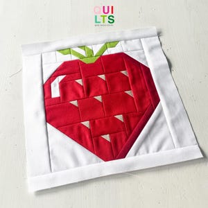 May include: A handmade quilt block with a red strawberry design, green leaves, and white accents. The strawberry is set against a white background. The text "QUILTS AND NICE STUFF" is visible in the upper right corner.