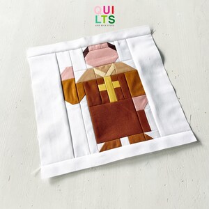Pode incluir: Um bloco de colcha de tecido quadrado com um design de peças de uma figura com uma túnica castanha e uma cruz amarela no peito. A figura tem rosto e mãos cor-de-rosa claro, com uma mão levantada. Enquadrado por uma borda branca.