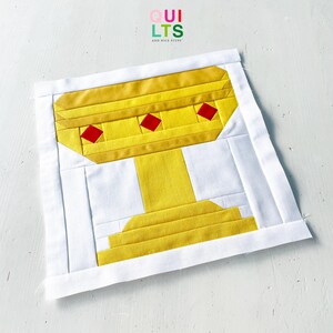May include: A square fabric quilt block featuring a pieced yellow chalice design on a white background. The chalice is accented with three small red diamond shapes across the top rim. The text QUILTS AND NICE STUFF is printed at the top center.