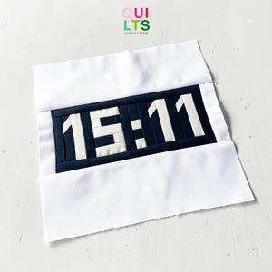 Peut inclure: Un bloc de courtepointe fait à la main représentant l'heure 15:11 en blanc sur un fond bleu marine. Les chiffres sont construits à partir de petits morceaux de tissu. Le bloc est encadré de tissu blanc, avec le texte "QUILTS AND NICE STUFF" en haut à droite.