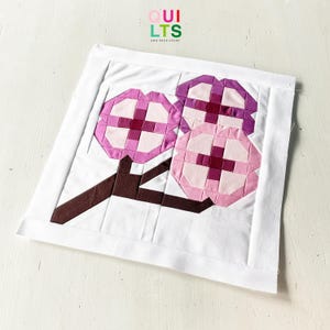 May include: A handmade quilt square with three stylised flower blossoms in pink and purple, with a brown stem, on a white background. The text "QUILTS AND NICE STUFF" is in the upper right corner.