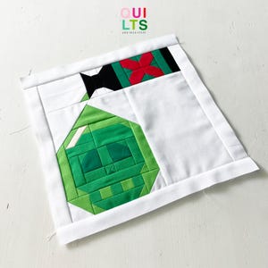 Könnte beinhalten: Ein handgefertigter Quiltblock mit einem geometrischen Design aus grünem, schwarzem, rotem und weißem Stoff. Der Block hat eine zentrale grüne Form, eine schwarze Form und ein rotes Kreuzdesign. Der Text "QUILTS AND NICE STUFF" befindet sich in der oberen rechten Ecke.