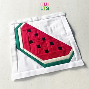 Könnte beinhalten: Ein handgefertigtes Quiltquadrat mit einem Wassermelonen-Scheiben-Design. Die Wassermelone hat ein rotes Inneres mit schwarzen quadratischen Samen, eine weiße Schale und eine grüne äußere Schale. Der Quilt ist vor einem weißen Hintergrund platziert, mit dem Text "QUILTS AND NICE STUFF".