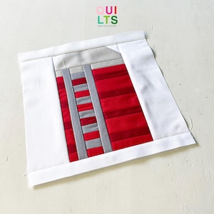 May include: A square fabric quilt block featuring a central design of horizontal red and dark red fabric strips intersected by two vertical grey fabric strips. The center is framed by a white border and a light grey top section, with the text QUILTS AND NICE STUFF printed above.