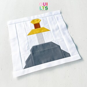 May include: A square fabric quilt block featuring a pieced design of a sword embedded in a stone. The sword has a yellow crossguard, a brown handle, and a light grey blade. The stone is dark grey, set against a white background with the text QUILTS AND NICE STUFF at the top.