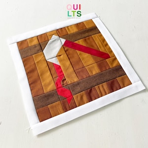 Könnte beinhalten: Ein handgefertigtes Quiltquadrat mit einem Drachendesign. Der Drachen ist weiß mit einem roten Schwanz, vor einem Hintergrund aus braunen und beigefarbenen Stoffblöcken. Das Quadrat ist mit einem weißen Rand versehen. Der Text "QUILTS AND NICE STUFF" befindet sich oben.