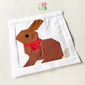 May include: A handmade quilt block depicting a brown rabbit with a red scarf on a white background. The block is framed with a white border, and the text "QUILTS AND NICE STUFF" is visible in the upper right corner.