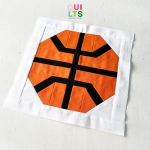 May include: A white fabric square with a black and orange basketball design in the center. The design is made of fabric squares sewn together to create the basketball pattern.