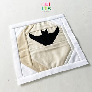 Könnte beinhalten: Ein handgefertigtes Quiltquadrat mit einem schwarzen Fledermaus-Design auf beigem Hintergrund, umrahmt von weißen und beigen geometrischen Formen. Das Quiltquadrat ist mit einem weißen Stoff eingefasst und der Text "QUILTS AND NICE STUFF" ist oben rechts sichtbar.